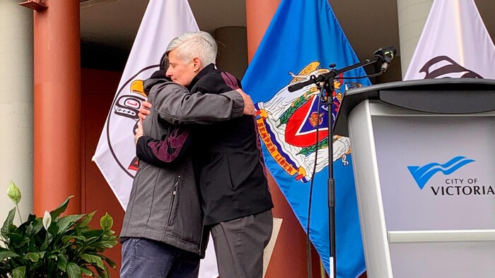 Le chef de la Première Nation Songhees, Ron Sam, et la mairesse de Victoria, Lisa Helps, s'enlacent devant des drapeaux.