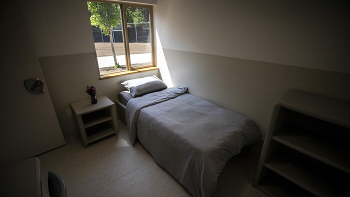 L'intérieur d'une chambre. Un lit sous une fenêtre avec une table de chevet, une étagère, un bureau et une chaise. Sur la table de chevet, il y a un pot avec des fleurs.