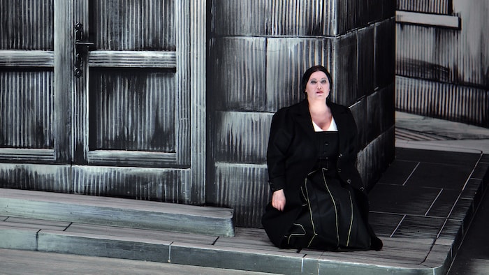 Une chanteuse d'opéra assise sur scène dans un décor composé de bois et de pierres factices. 
