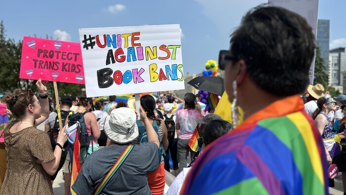 Une personne vêtue aux couleurs de l'arc-en-ciel regarde deux personnes de dos qui tiennent des pancartes dans un défilé de la Fierté à Winnipeg en juin. Sur les pancartes, on peut lire «Protégez les enfants transgenres» et «Unissons-nous contre l'interdiction des livres».