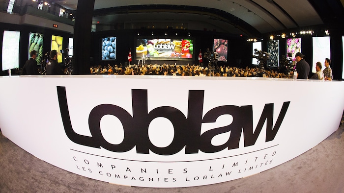 Sur une photo de type œil de poisson, le logo de Loblaw apparaît à l'entrée d'une salle de conférence remplie de gens.