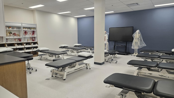 Une salle de classe de physiothérapie avec des tables de traitement, des modèles anatomiques et un grand écran noir.