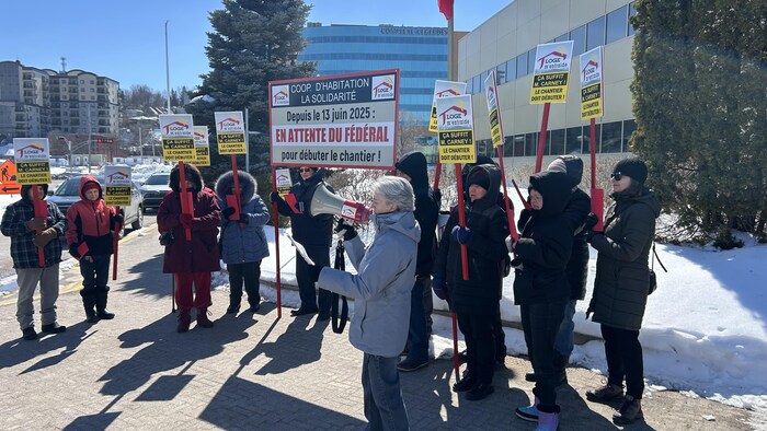 Des personnes tenant des pancartes sont debout en demi-cercle autour d'une femme tenant un mégaphone.