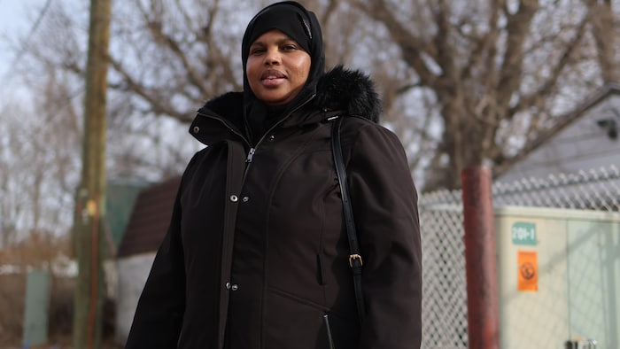 Une femme portant un manteau d'hiver foncé et un hijab noir se tient à l'extérieur devant des arbres dénudés.