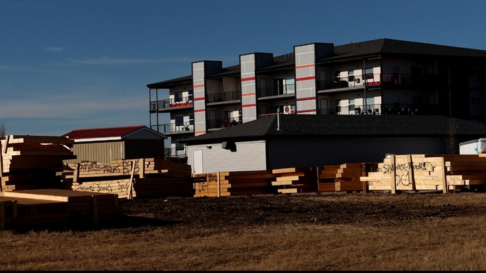 Des piles de bois d'œuvre sont entreposées devant un immeuble d'habitation moderne sous un ciel bleu.