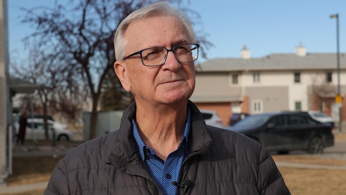 Un homme à lunettes portant un manteau matelassé regarde vers le haut dans un quartier résidentiel.