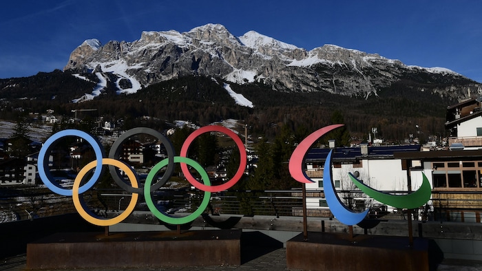 Le logo des Jeux olympiques et paralympiques de Milan-Cortina avec, en arrière-plan, une piste de ski alpin.
