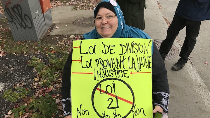 Une femme porte le voile et un chapelet en tenant une affiche qui dénonce la loi 21.