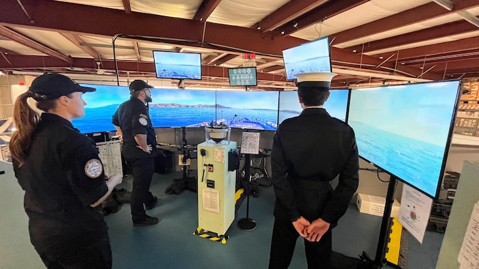 Trois membres de la Marine utilisent un simulateur de navigation. La vue montre trois écrans d'ordinateur, un pont de navire, de l'équipement et des moniteurs qui affichent un océan et un quai.