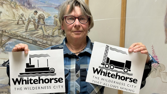 Une femme tient deux images dans ses mains. À gauche, le logo officiel de la Ville de Whitehorse, avec un bateau. À droite, un nouveau logo si la ville allait de l'avant avec l'exploration minière, dans lequel le bateau est remplacé par un camion.