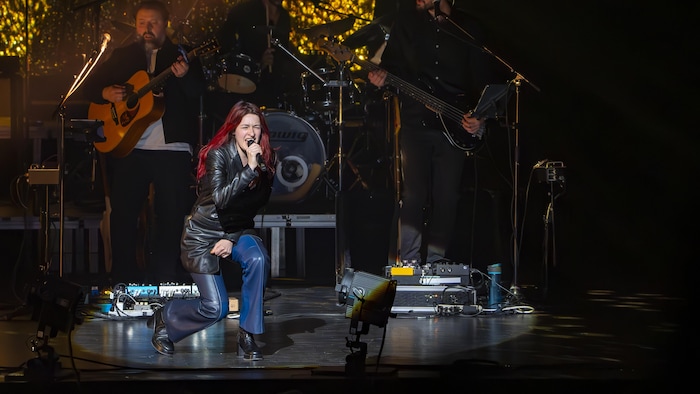 Lou-Adriane Cassidy, avec ses longs cheveux rouges, chante sur scène, un genou fléchit.