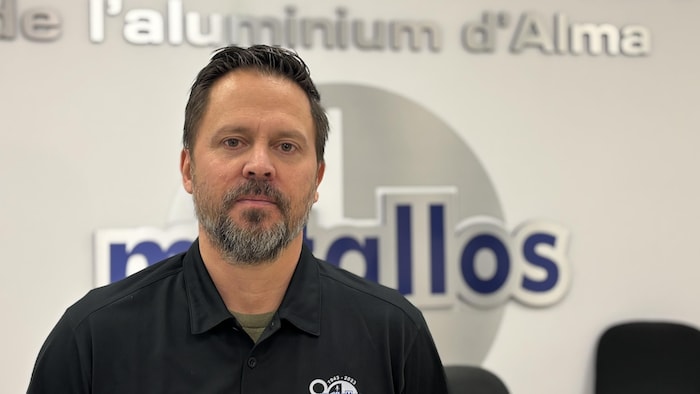 Un homme d'âge moyen avec une barbe courte, vêtu d'un polo noir arborant un logo, pose devant un mur blanc. En arrière-plan, on aperçoit l'inscription « Syndicat des travailleurs de l'aluminium d'Alma » et le logo des Métallos.