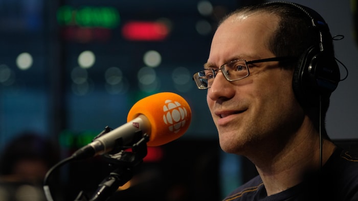 Un homme portant des lunettes rectangulaires, dans un studio de radio derrière un micro.