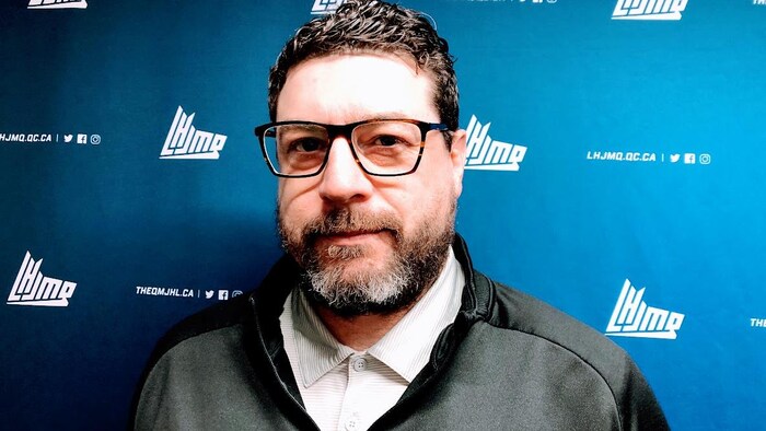 Un homme portant des lunettes debout devant un panneau bleu décoré de logos de la LHJMQ.