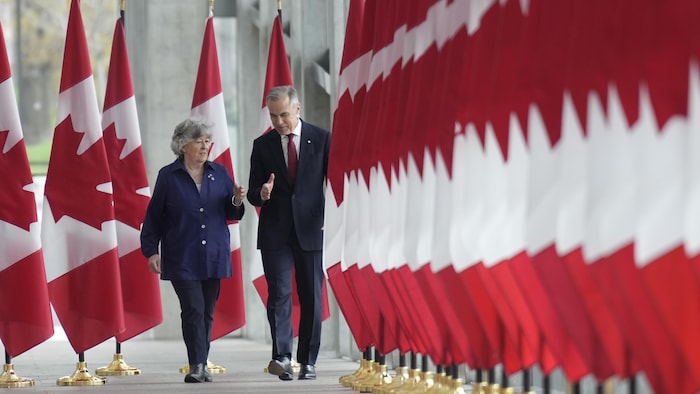 Louise Arbour et Mark Carney marchent entre des drapeaux du Canada.