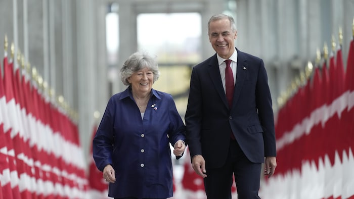Mark Carney et Louise Arbour marchent dans un couloir bordé de drapeaux canadiens.