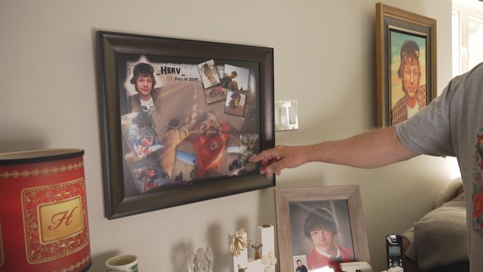 Un homme nous pointe un tableau où on voit des photos de son fils décédé