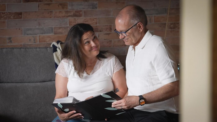 Caroline Lévesque et Luc Berthold tiennent un album photo, assis sur un divan.
