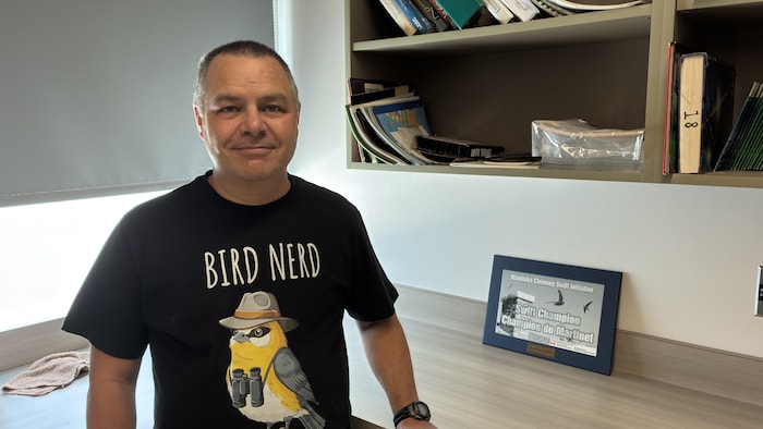 Luc Blanchette, enseignant à l'École de la région Saint-Jean-Baptiste et ornithologue amateur, se tient debout dans une salle de classe, avec un t-shirt sur lequel est écrit "Bird Nerd"