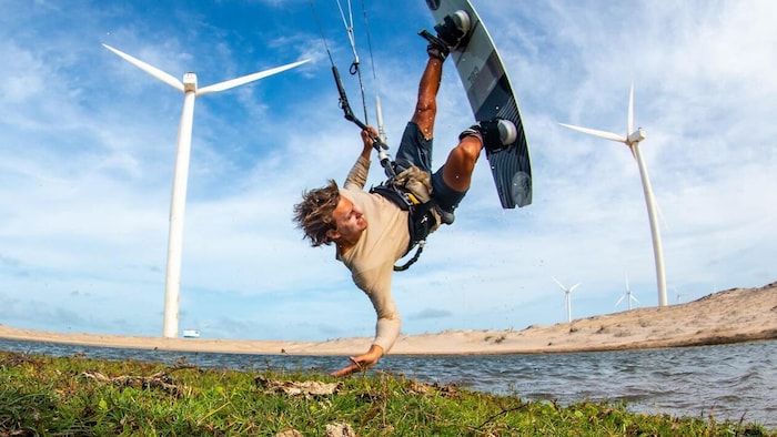 Lucas Arsenault fait du kite surf. Il a la tête à l’envers, devant un paysage estival où l’on aperçoit un cours d’eau et des éoliennes. 