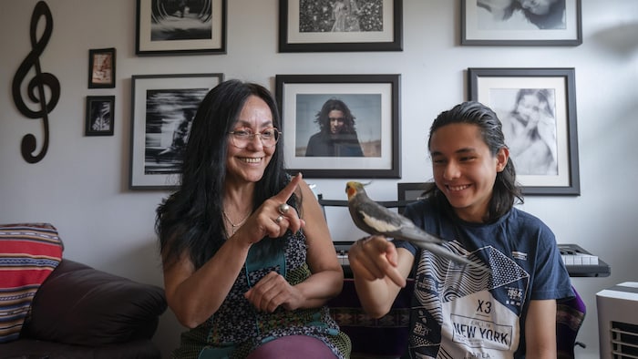 Une mère et son fils adolescent, qui a un oiseau perché sur le doigt, sourient en regardant l'oiseau, dans leur appartement.