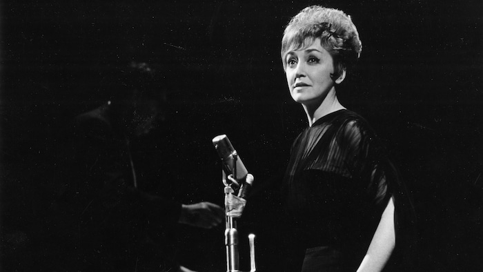 Lucille Dumont sur scène, devant un micro. Derrière elle on aperçoit, dans
l'ombre, un chef d'orchestre non identifié.