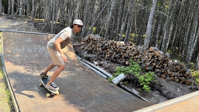 Un homme en short et t-shirt fait de la planche à roulettes sur une rampe en bois courbée au milieu d'une zone boisée à côté d'une pile de cordes de bois.