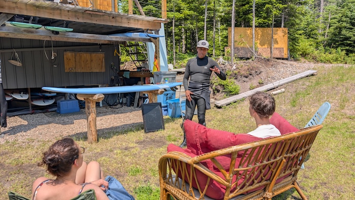 Un homme en combinaison s'adresse à deux personnes assises à l'extérieur devant un bâtiment en bois équipé de supports pour planches de surf.