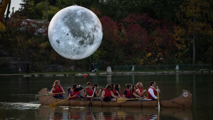 Plusieurs personnes dans une barque sur un lac, devant une réplique de la lune. 