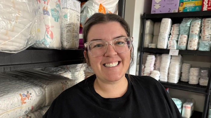Une femme souriante qui porte des lunettes à grosse monture transparente est adossée à l'intersection de deux hautes étagères remplies de couches pour bébés.