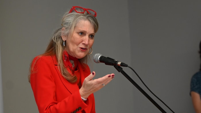 Une femme avec des lunettes à monture rouge posées sur le dessus de sa tête parle, debout devant un micro, la main droite levée et la paume vers le haut.