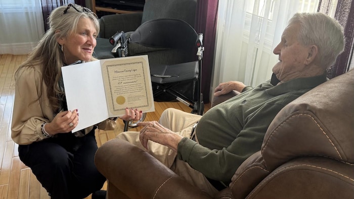 Lyne Chantal Boudreau agenouillé devant Emery Léger qui est dans son divan. Elle lui montre un certificat de reconnaissance.