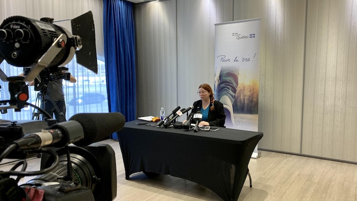La coroner Sophie Régnière, une femme aux cheveux roux et lunettes, est assise derrière une table noire recouverte de micros lors d'un point de presse. Elle est éclairée par des lumières de cinéma, avec plusieurs caméras et techniciens visibles au premier plan, accentuant l'angle de la caméra. Derrière elle se trouve une bannière verticale du Bureau du coroner du Québec avec la devise « Pour la vie ! ».