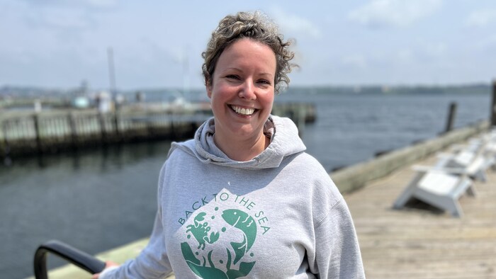Une femme fait un grand sourire, debout sur le front de mer, cheveux au vent et une main sur une rampe en métal