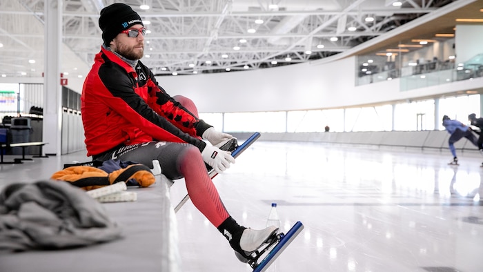 Laurent Dubreuil, assis au bord d'une patinoire intérieure, ajuste ses patins de vitesse tout en observant la glace.