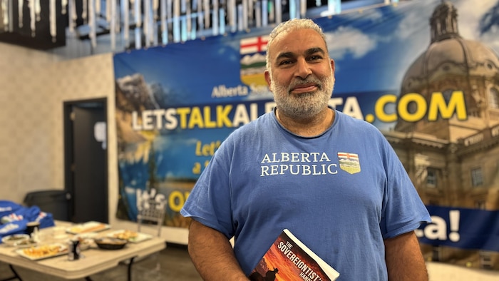 Un homme avec un t-shirt bleu pose devant une affiche de Let's Talk Alberta.