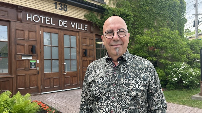 Un homme chauve avec des lunettes et une barbichette sourit et regarde la caméra. Il porte une chemise à motifs sombres. Derrière lui se trouve un bâtiment en briques avec "HOTEL DE VILLE" inscrit au-dessus d'une grande porte en bois.