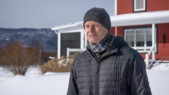 L'homme regarde vers l'horizon. Derrière lui, il y a une maison et un paysage gaspésien en hiver.