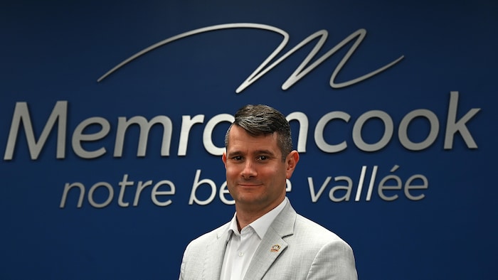 Le maire de Memramcook, Maxime O. Bourgeois, vêtu d'un veston clair, devant un mur bleu affichant le logo de la municipalité.