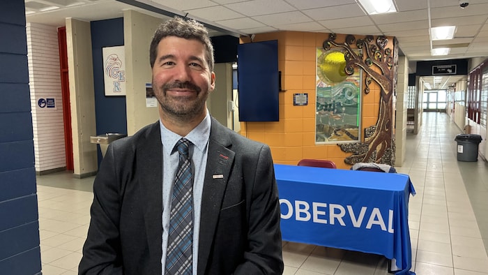 Un homme souriant portant un veston sombre, une chemise bleue et une cravate se tient au premier plan dans un couloir d'établissement scolaire.