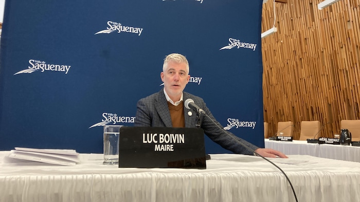 L'image montre Luc Boivin, maire de la ville de Saguenay, assis derrière un micro lors d'une séance officielle. Il est devant un rideau bleu arborant le logo de la ville. Des documents et un pichet d'eau sont sur la table.
