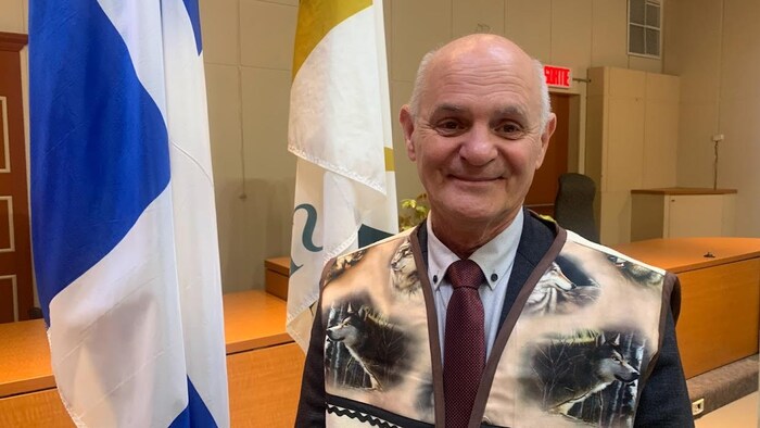 Denis Miousse porte une veste offerte par le chef de la communauté innue, il sourit devant les drapeaux officiels. 