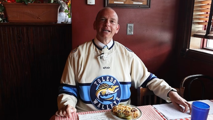 Wade Kapszukiewicz assis à une table d'un restaurant avec un hot dog devant lui dans son assiette et un chandail du Walleye de Toledo.
