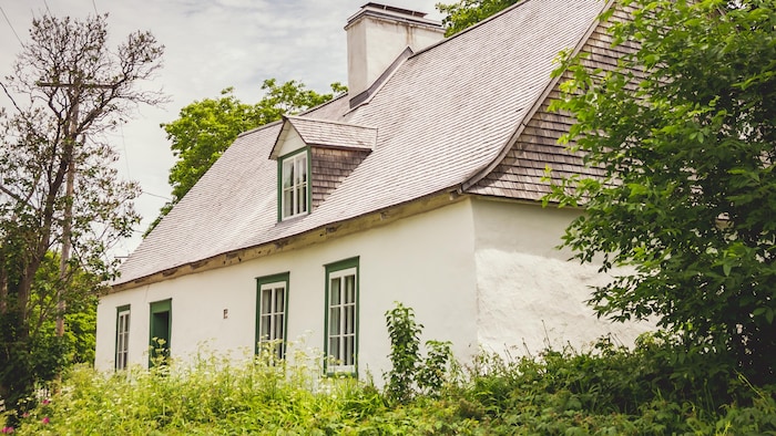 Maison ancestrale au toit de bardeaux, entourée d’une végétation dense sous un ciel nuageux.