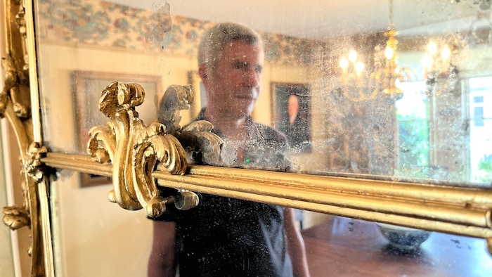 Un homme regarde son reflet terni dans un vieux miroir de style rococo.