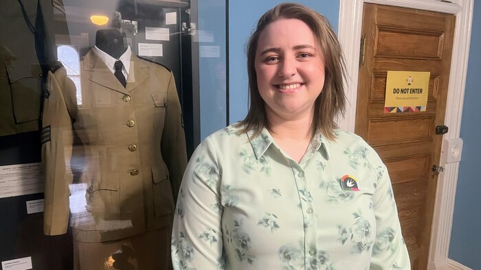 Une femme souriante pose devant une vitrine de musée contenant un uniforme militaire.