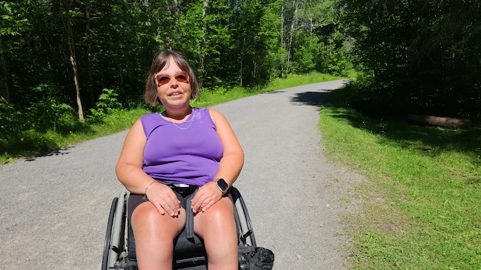 Une femme portant un haut violet et des lunettes de soleil sourit, assise dans un fauteuil roulant au premier plan. Elle se trouve sur un chemin de gravier ombragé qui serpente à travers une forêt verdoyante par temps ensoleillé.