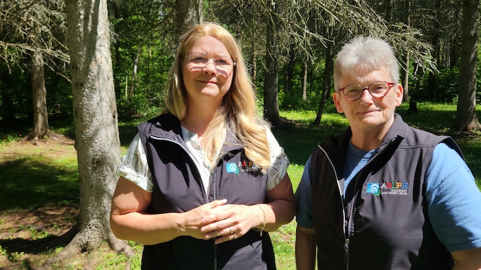 Deux femmes portant des lunettes et des vestes sans manches identiques, ornées d'un logo, posent côte à côte à l'extérieur. Elles se tiennent debout dans une forêt ensoleillée, entourées de grands arbres et d'une pelouse verdoyante.