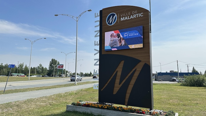L'affiche de bienvenue de Malartic, un grand panneau en bois monté sur un petit bac à fleurs, à l'entrée de la ville.