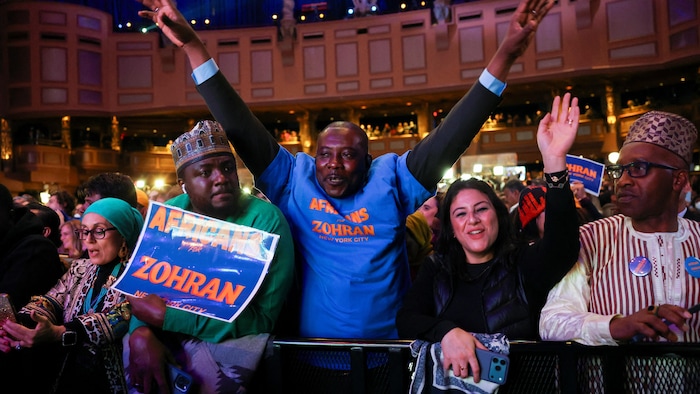 De nombreux partisans rassemblés dans une salle avec des affiches et des t-shirts de Zohran Mamdani célèbrent, souriants, certains en levant les bras.